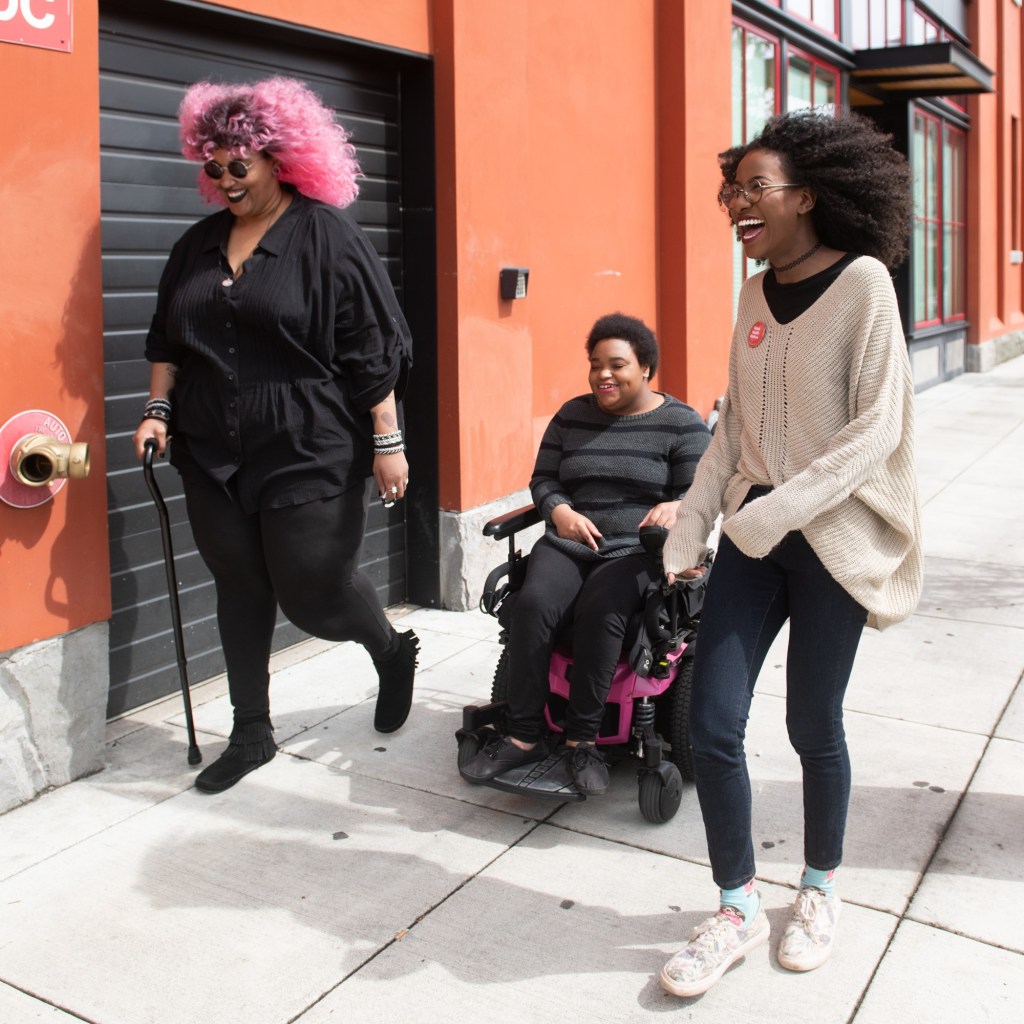Three Black and disabled folx laugh uproariously while strolling down a sidewalk on a windy day. On the left, a non-binary person walks with a cane in one hand and a tangle stim toy in the other. In the middle, a non-binary person rolls along in their power wheelchair. On the right, a femme walks with fabulously windswept hair. A street parking meter is in the background on the right.